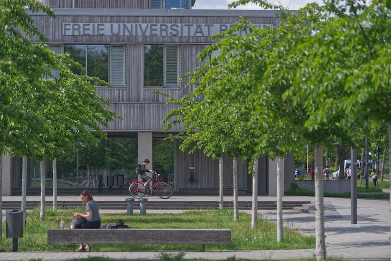 Outside view of Freie Universität Berlin. Grey colour building is behind 8 trees and one bike is moving in front. A girl is sitting on the bench outside the university
