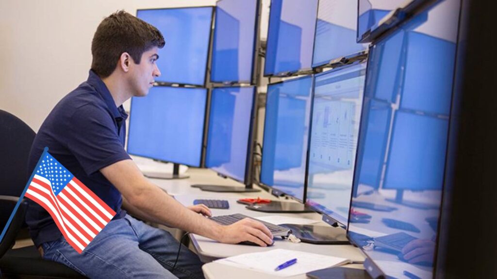 PhD in Computer Science in USA - A student is stitting in front of 10 computer screens and working. There is a flag of USA on bottom left corner.